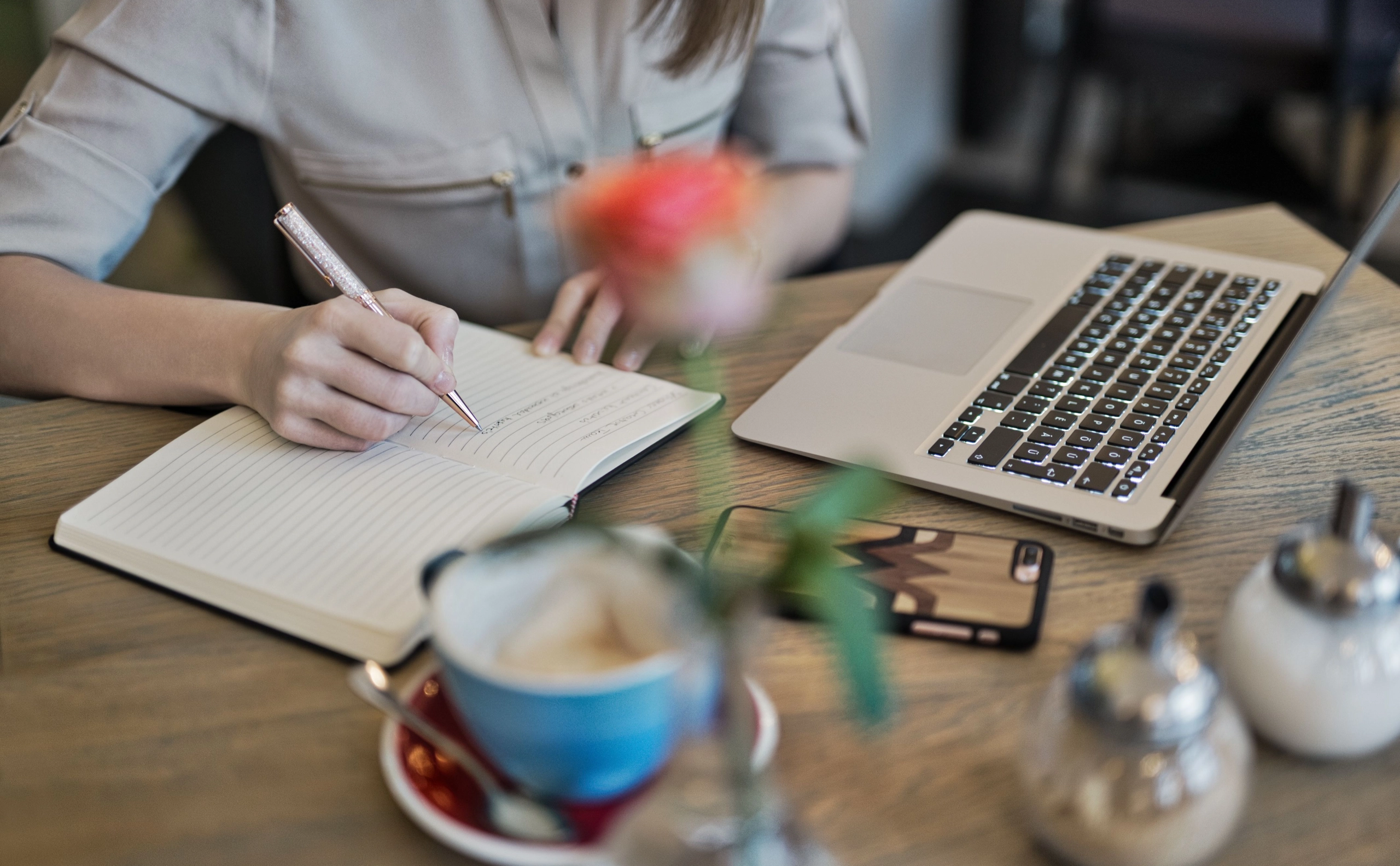 Persona escribe notas para un artículo en una libreta junto a su laptop y un café.